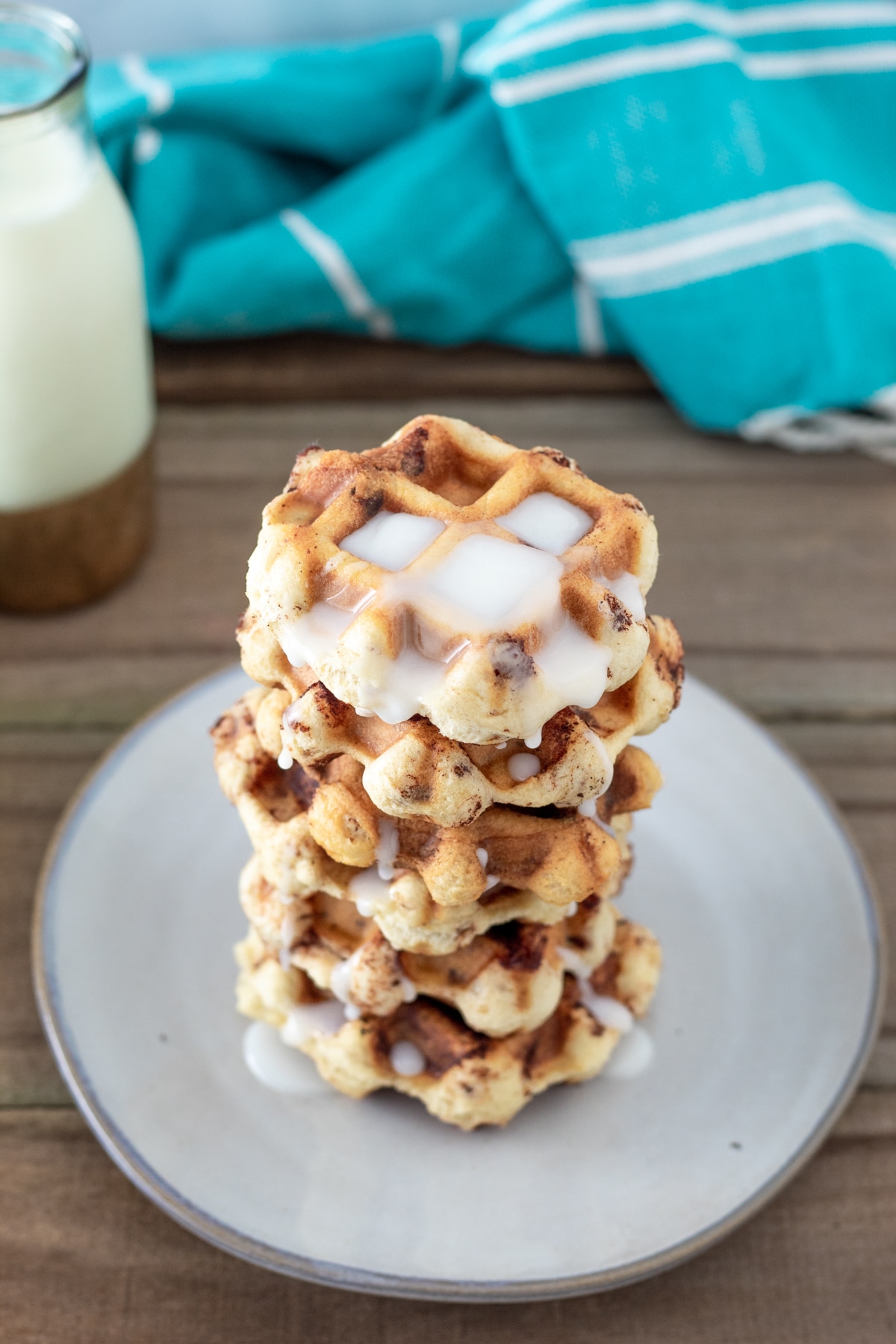 A white plate stacked with Cinnamon Roll Waffles topped with icing. A blue towel and a glass of milk are in the background