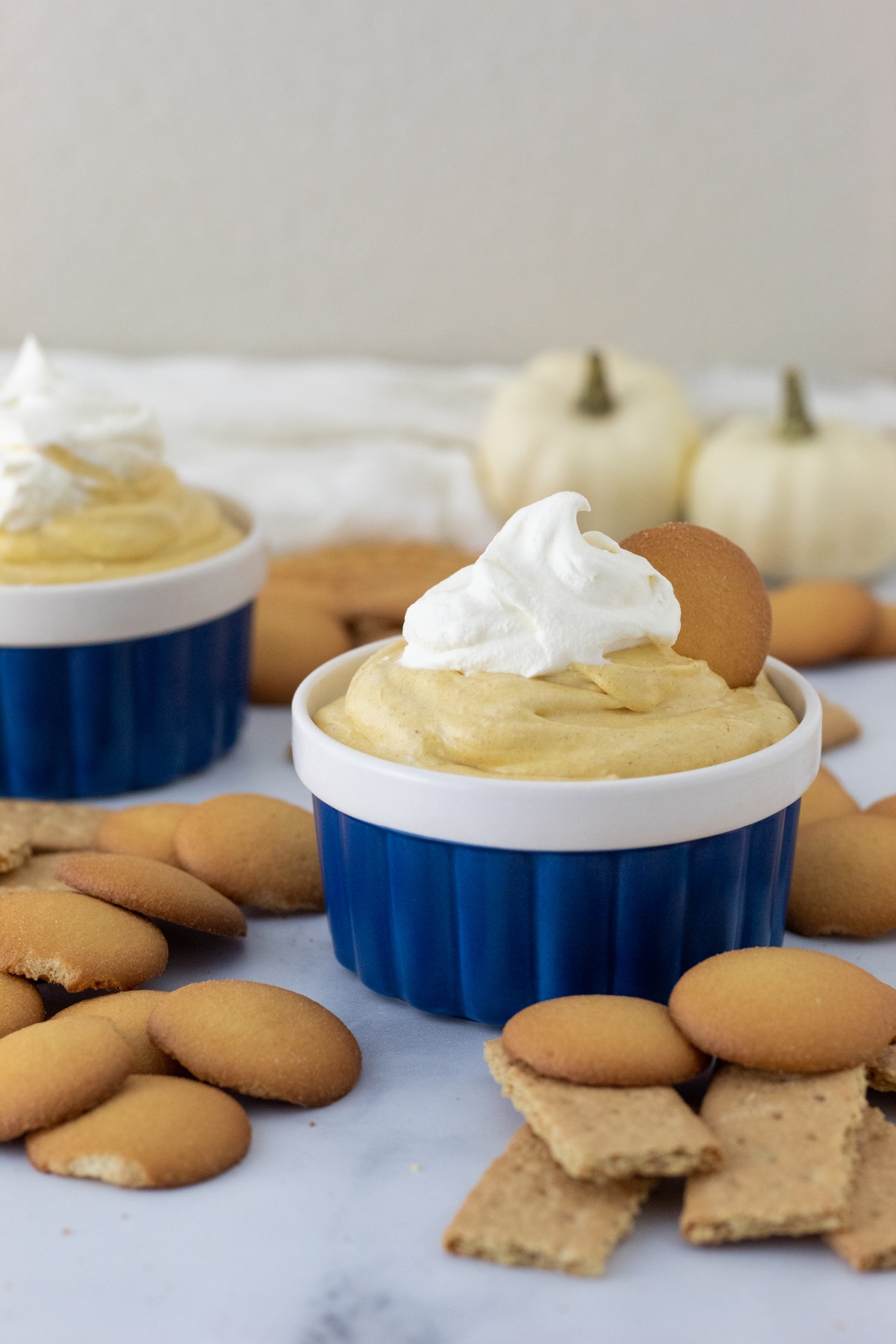 Two small blue bowls filled with Pumpkin Pie Dip topped with whipped cream. Nilla Wafers and Graham Crackers placed around bowls. Two small white pumpkins in background