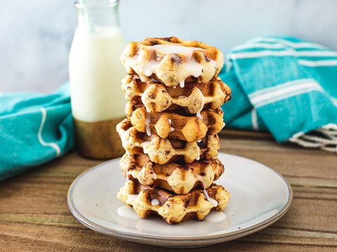 A stack of cinnamon roll waffles sitting on a plate with a cup of milk in the background